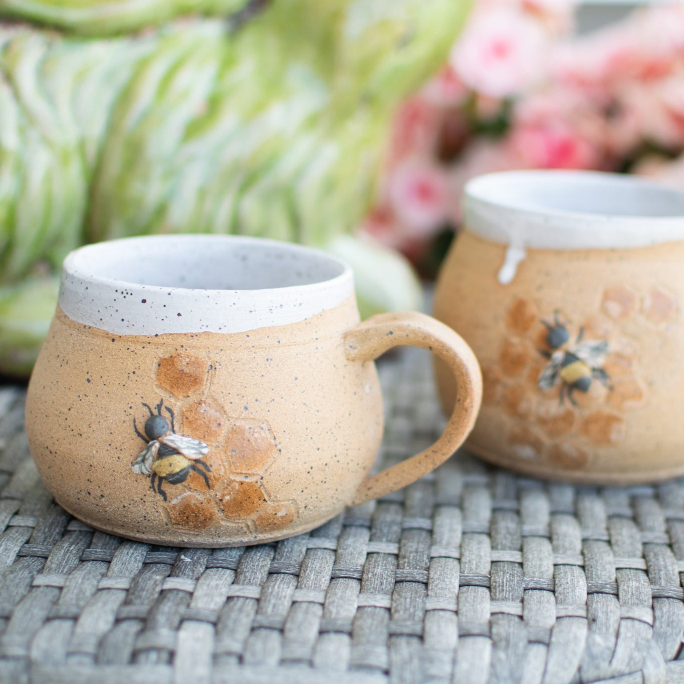Two ceramic mugs with bee designs on a woven surface.