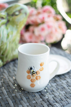 White mug with a bee design on a woven surface with flowers in the background