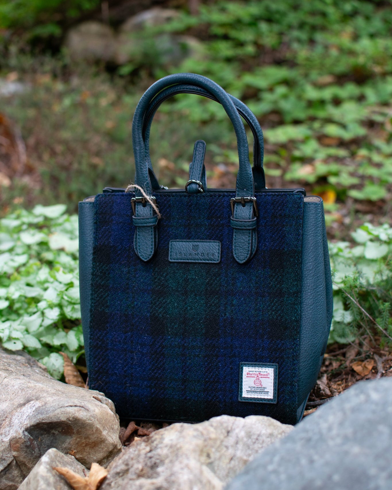Blue plaid handbag with a visible brand label on a natural background of rocks and plants.