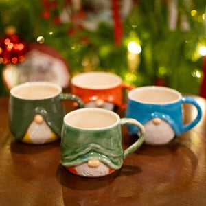 Four gnome-shaped mugs on a wooden table with a Christmas tree in the background.
